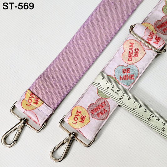 a pink strap with heart-shaped embroidered phrases such as "LOVE ME", "SWEET PEA", and "DREAM BIG". The strap is attached to a metal clasp, and there is a ruler placed next to it, indicating the strap's length.
