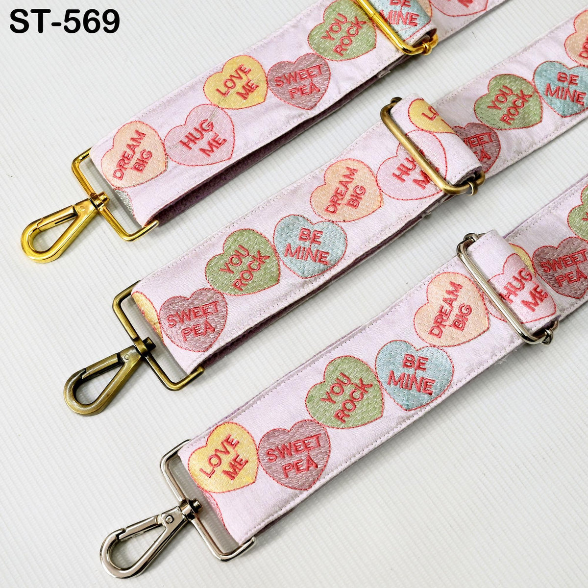a pink strap with heart-shaped embroidered phrases such as "LOVE ME", "HUG ME", "SWEET PEA", "BE MINE", and "DREAM BIG". The strap has a gold buckle and is displayed on a white background.