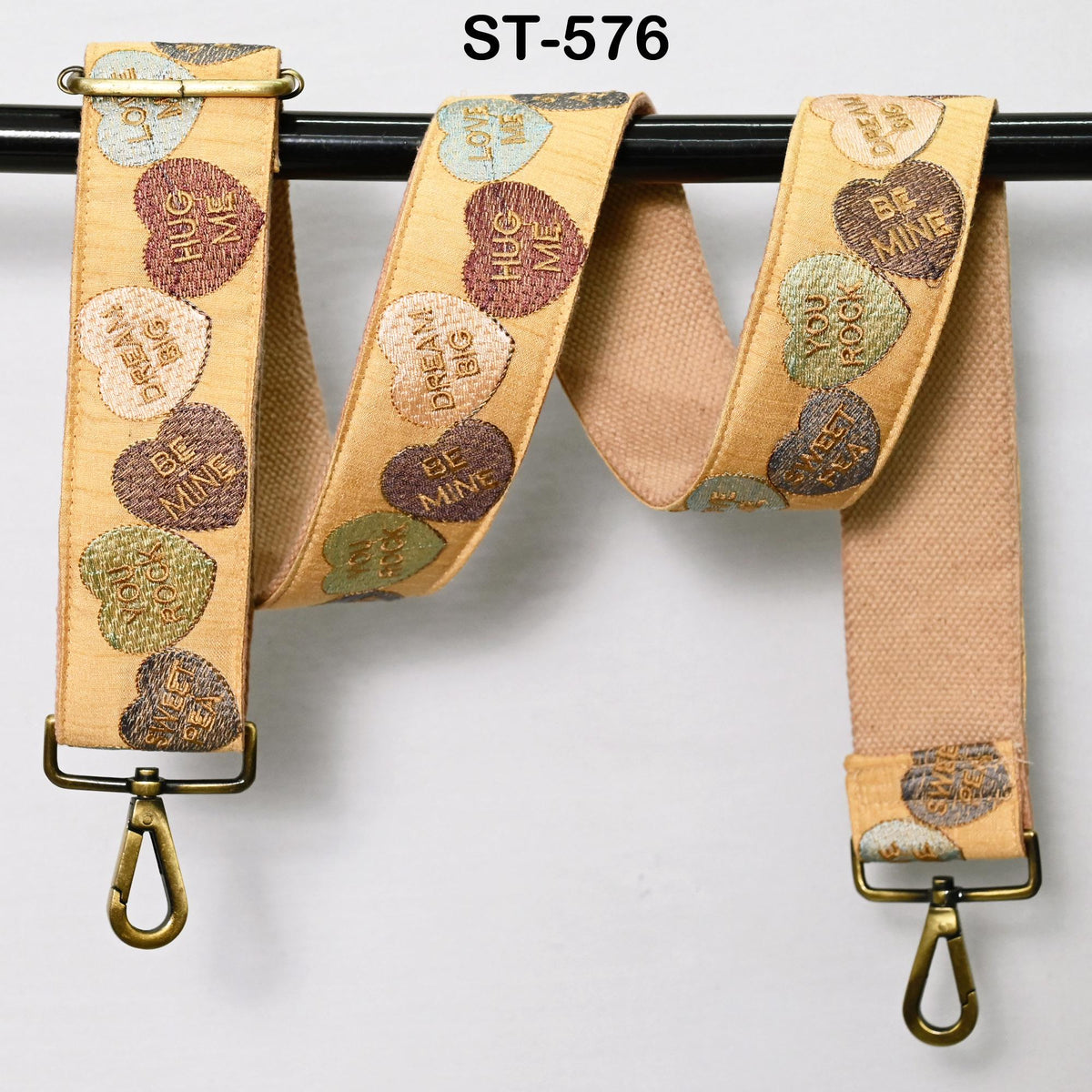 A beige strap with heart-shaped embroidered phrases such as "HUG ME", "DREAM BIG", "BE MINE", and "YOU LOVE ME" printed on it. The strap is hanging from a black hook or loop.