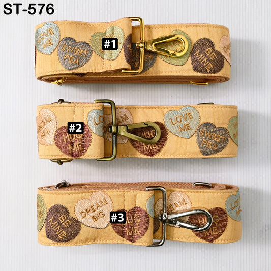 three beige straps with heart-shaped embroidered phrases on them. The straps have metal buckles and buckles on the front.