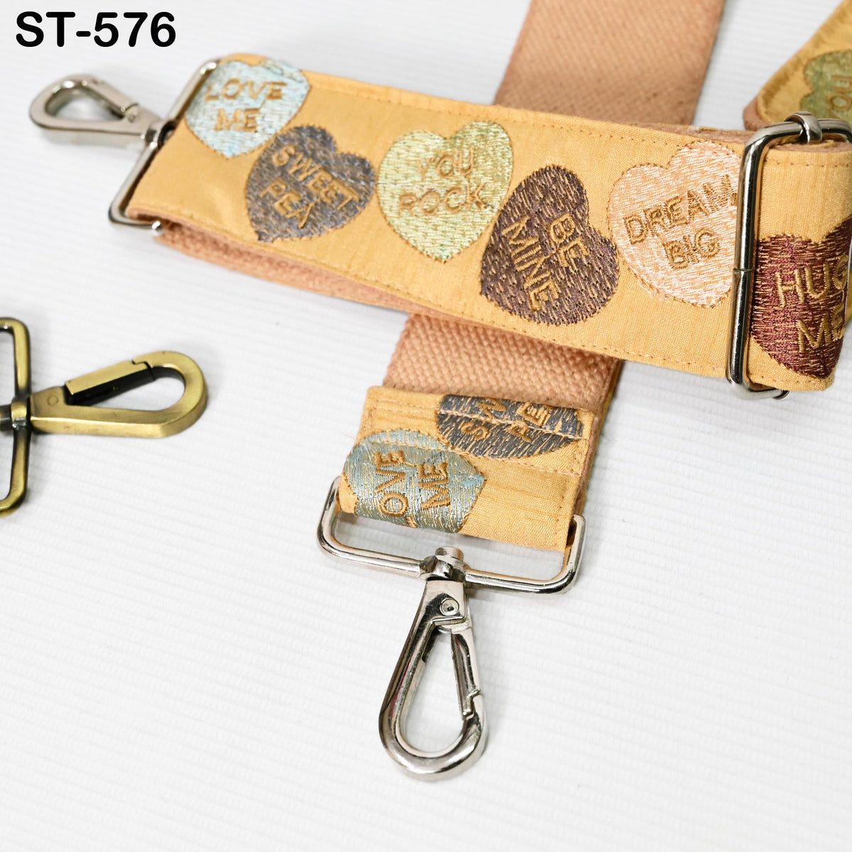 a beige strap with heart-shaped embroidered phrases such as "LOVE ME", "HUG ME", "DREAM BIG", and "SWEET PEAS", as well as a metal hook or clasp.