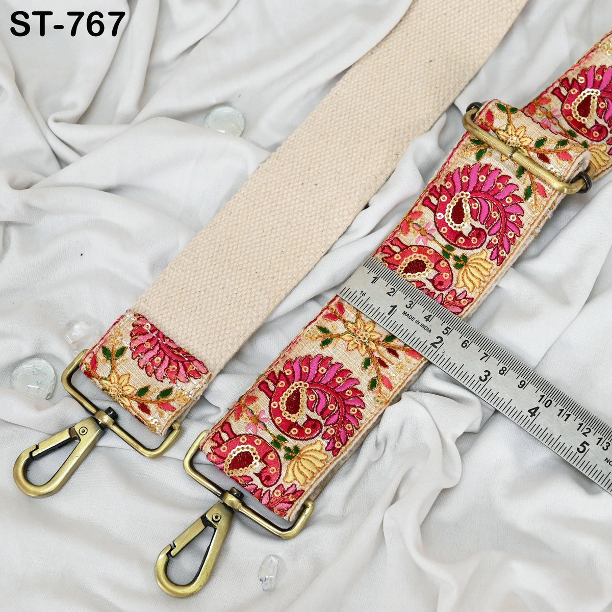 a beige strap with a floral pattern, resting on a white fabric surface. The strap has a metal clasp and a metal hook, and there is a ruler placed next to it.