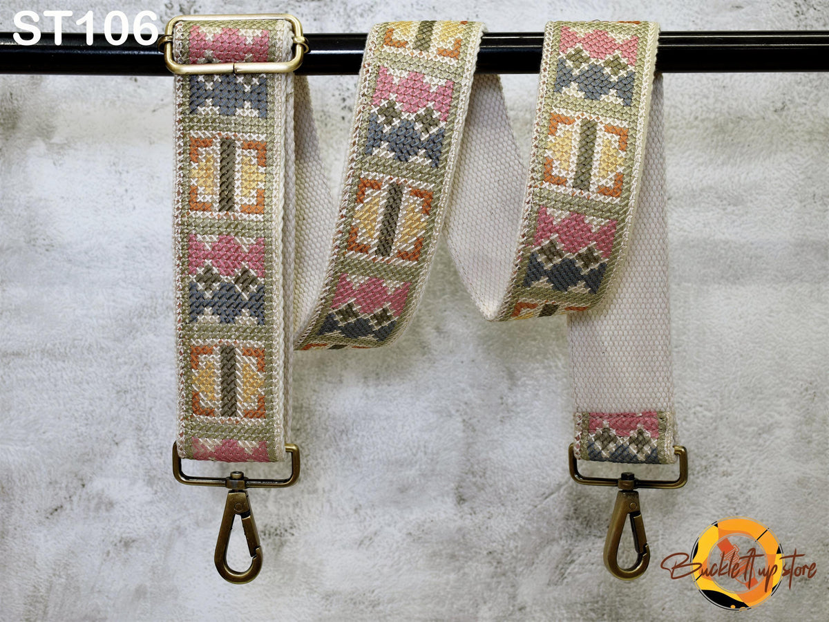 a colorful, patterned strap with a metal buckle attached to a black metal hook. The strap appears to be a cross-stitch or embroidered design, with a variety of colors and shapes. The strap is hanging on a black metal bar or hook, and there is a small logo or text in the bottom right corner of the image.