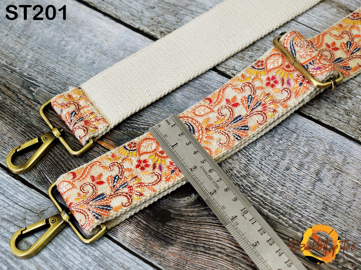 a white strap with a floral pattern and two gold metal buckles, placed on a wooden surface. There is a ruler placed next to the strap, indicating the strap's length.