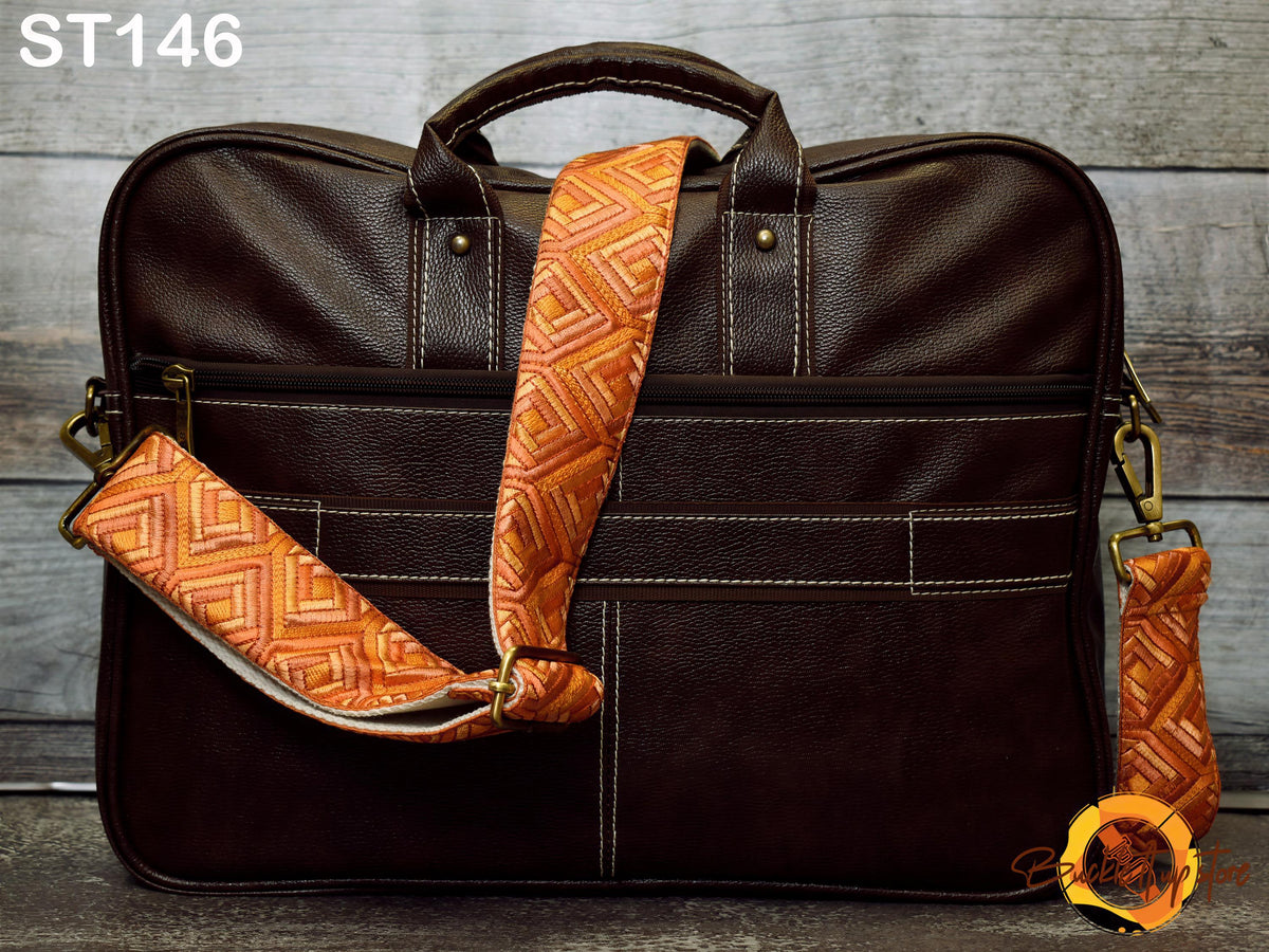 A brown leather laptop bag with a patterned orange strap, placed on a wooden surface against a wooden background.