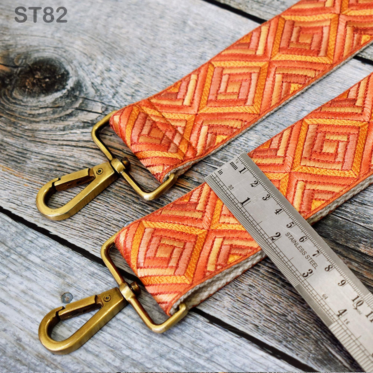 a pair of orange and gold patterned straps with metal clasps, resting on a wooden surface. A ruler is placed next to the straps, indicating the size or length of the straps.