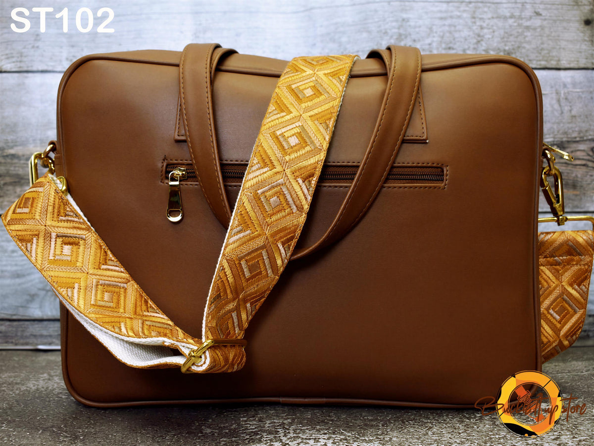 A brown leather laptop bag with a gold geometric patterned strap, placed on a wooden surface.