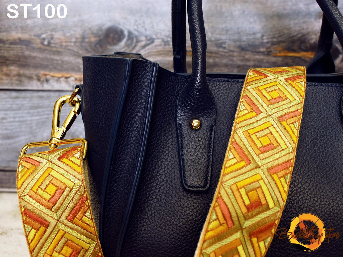 A close-up view of a black leather handbag with a gold-colored geometric patterned strap, placed on a wooden surface.