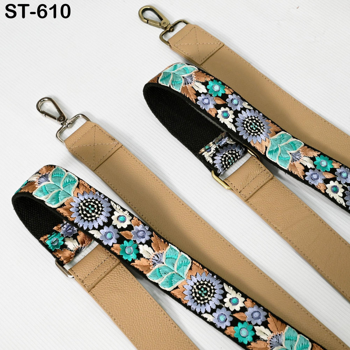 two beige straps with floral embroidery patterns, one strap is attached to a bag and the other is attached to a surface.