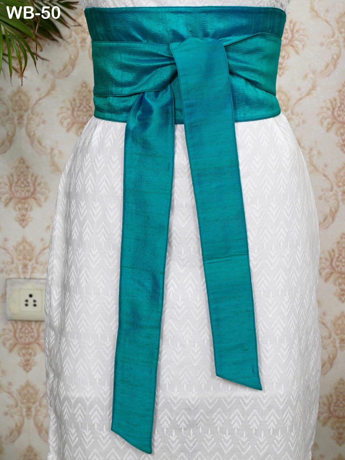 A turquoise sash tied around a white dress, displayed against a patterned wall.
