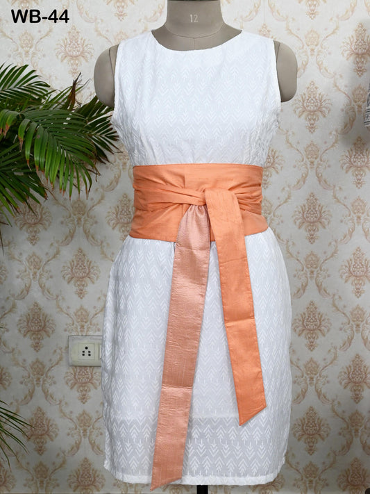A white dress with an orange sash, displayed on a mannequin against a patterned wallpaper.