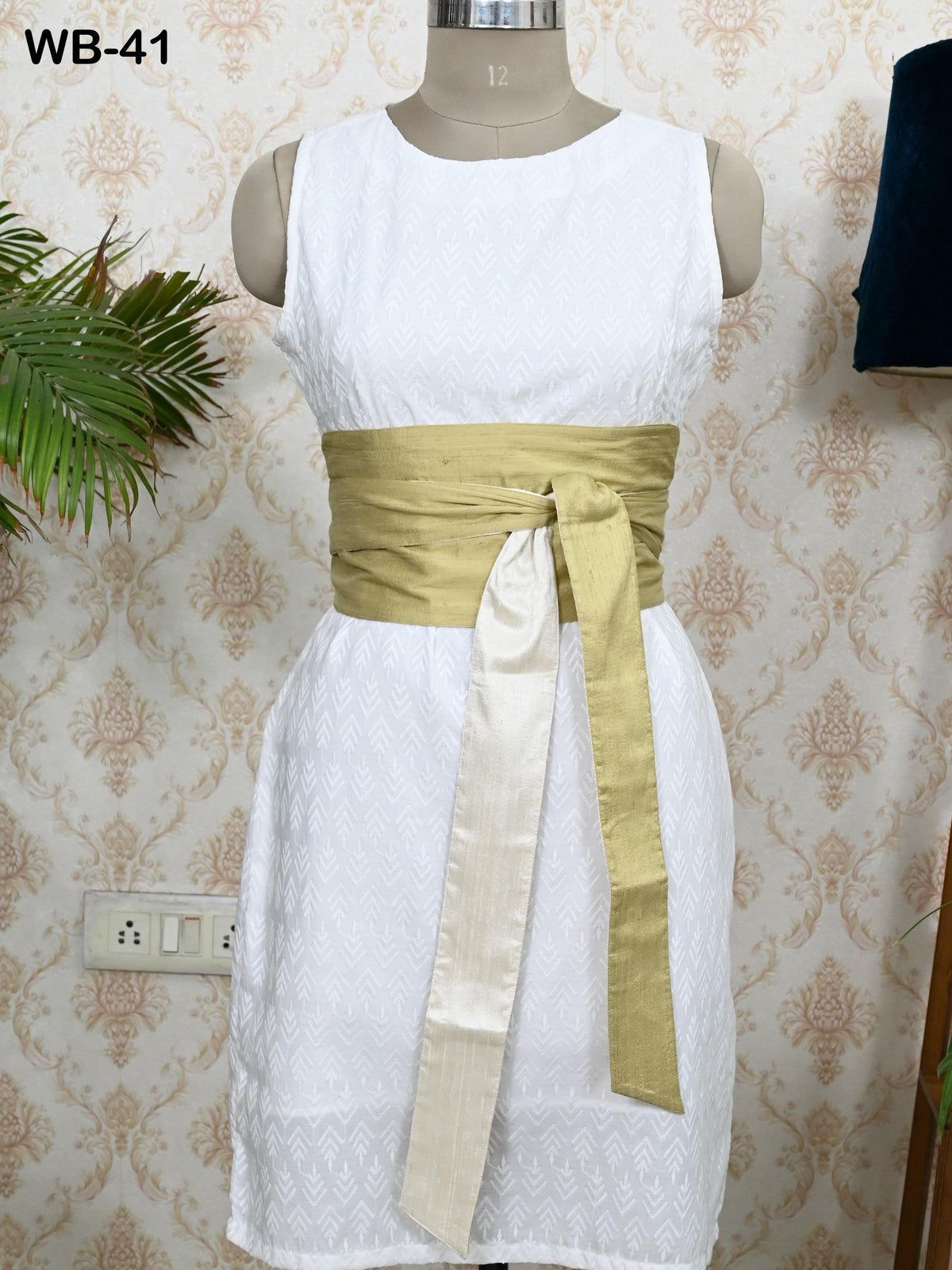 A white sleeveless dress with a gold sash tied around the waist, displayed on a mannequin against a patterned wallpaper.