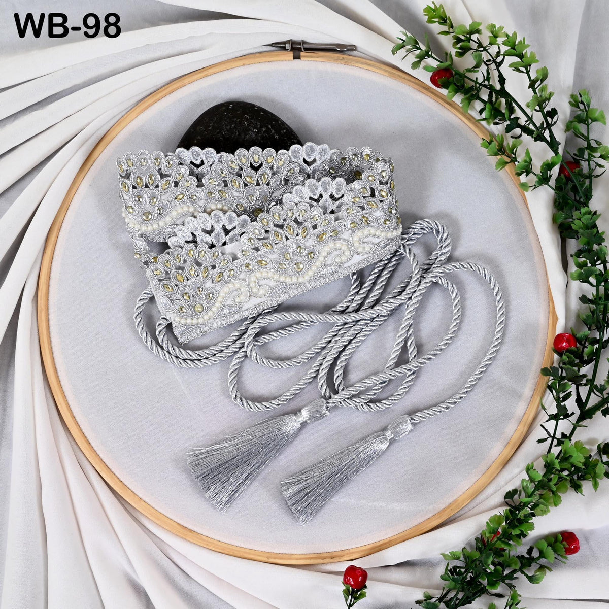 a white embroidery hoop with a decorative beaded design, a black stone, and a tassel with a fringe. The objects are arranged on a white background with greenery and red berries.