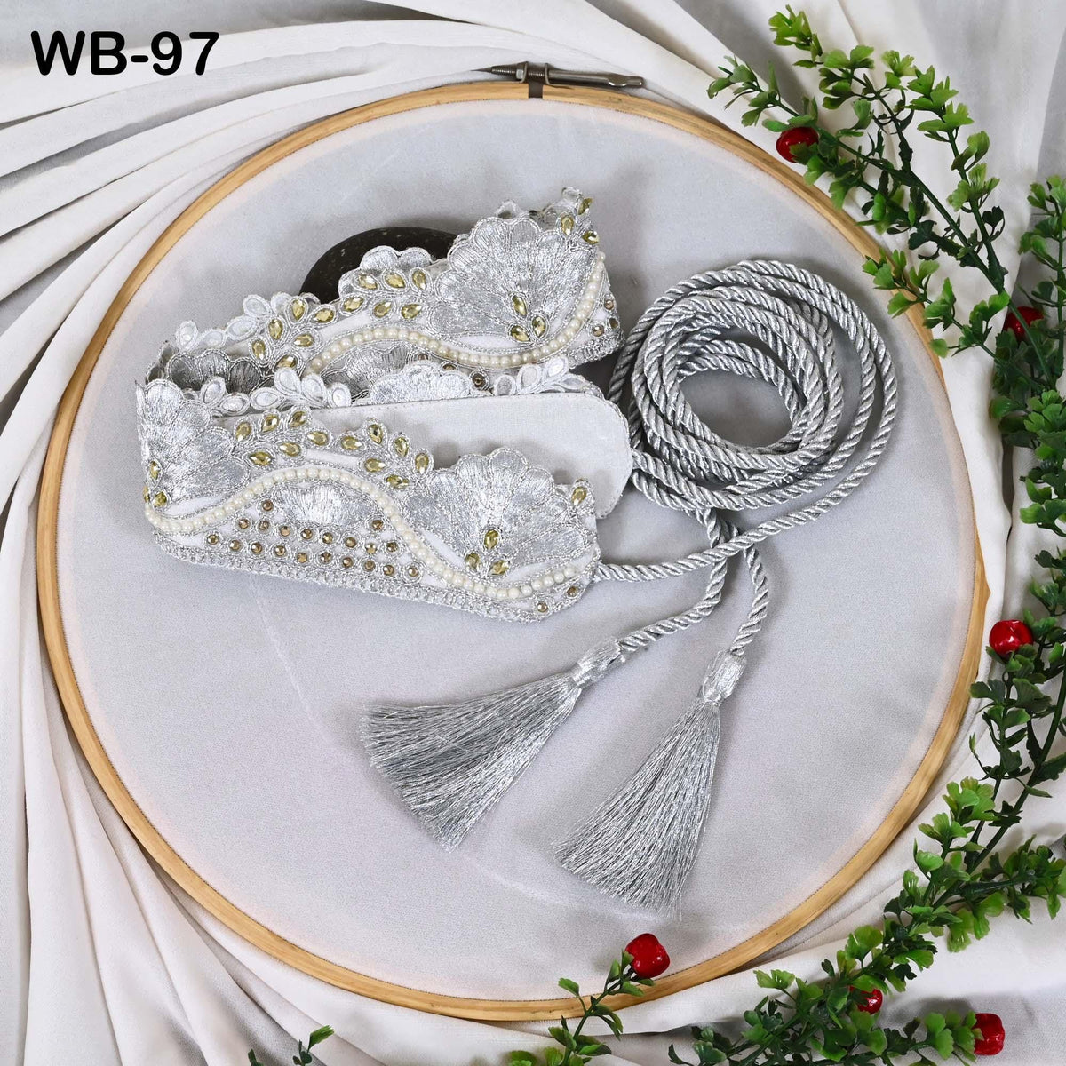 a white embroidery hoop with a silver and white lace-like design, a silver tassel, and a silver tangle cord. The background is white with greenery and red berries.