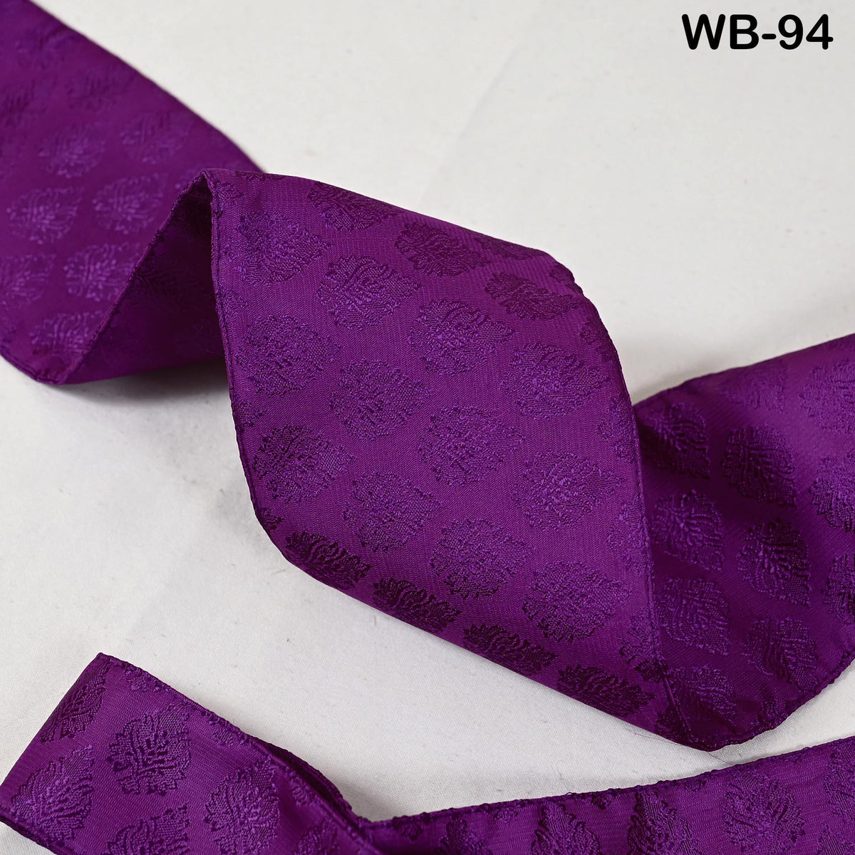 a close-up view of a purple fabric with a repeating pattern of small flowers or leaves. The fabric appears to be a type of ribbon or decorative material, possibly used for embellishment or as a decorative accent.