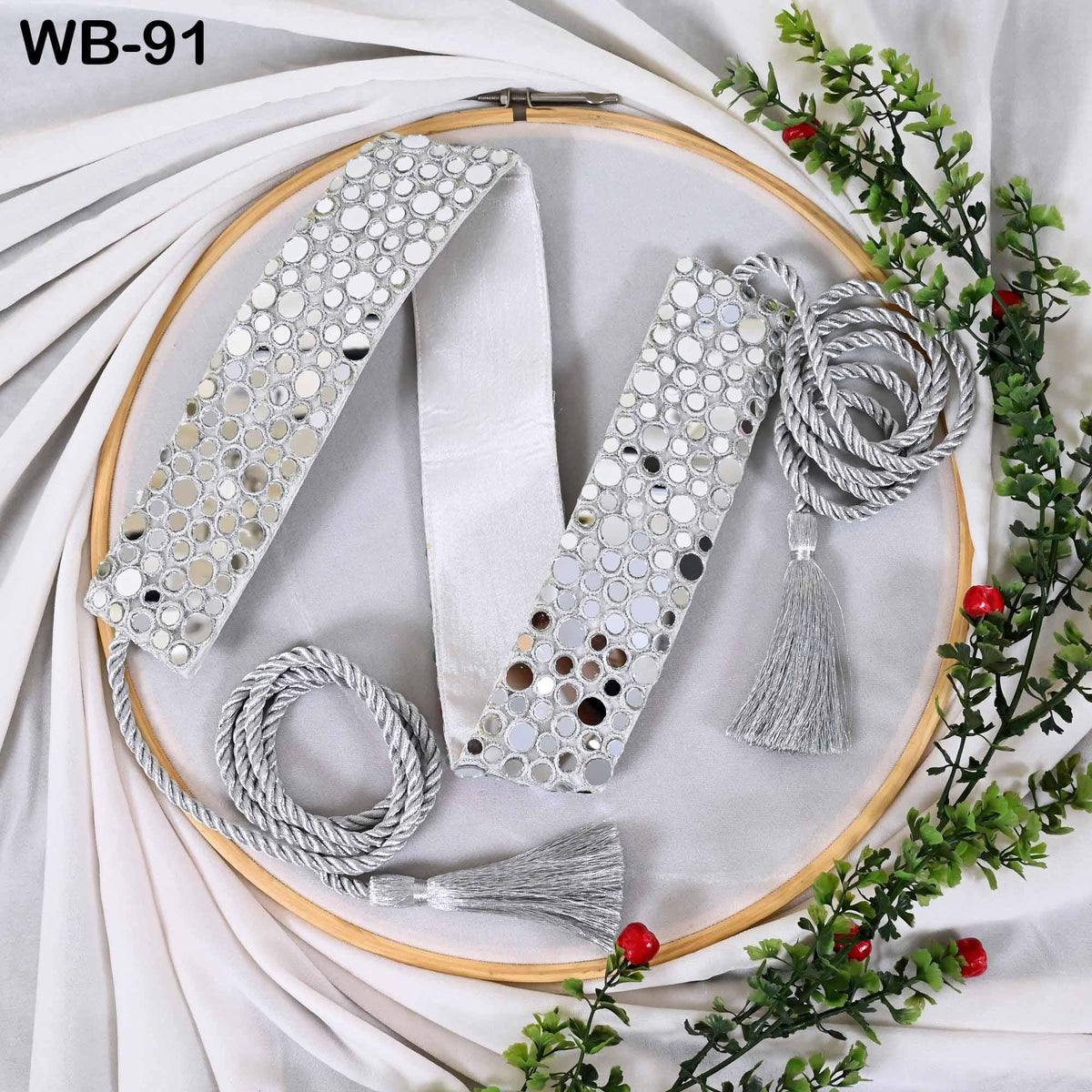 a silver-colored decorative item with a circular pattern, placed on a white surface. The item has a tassel and a ribbon attached to it, and there are some red berries and greenery surrounding the item.