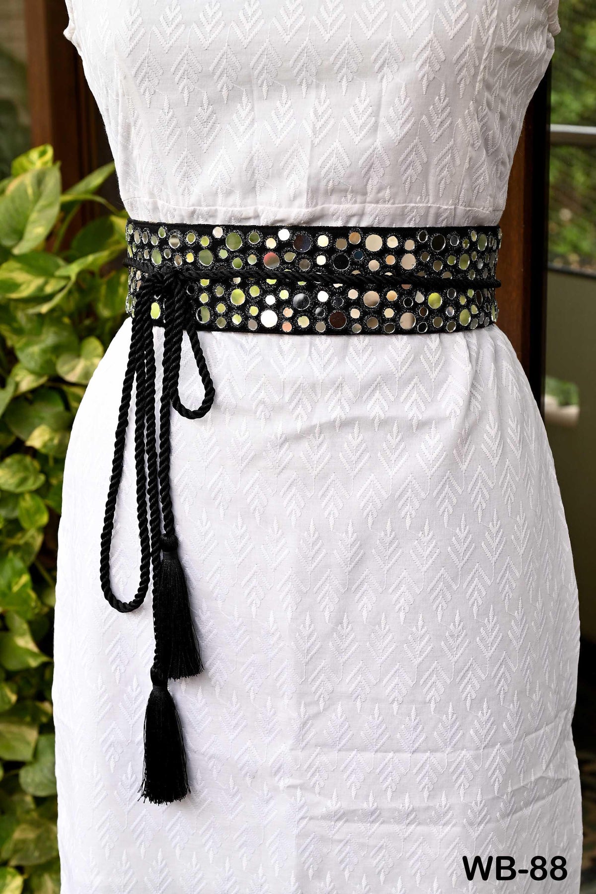 A white dress with a black tassel belt featuring small mirrors or circles on it, displayed in front of a plant.