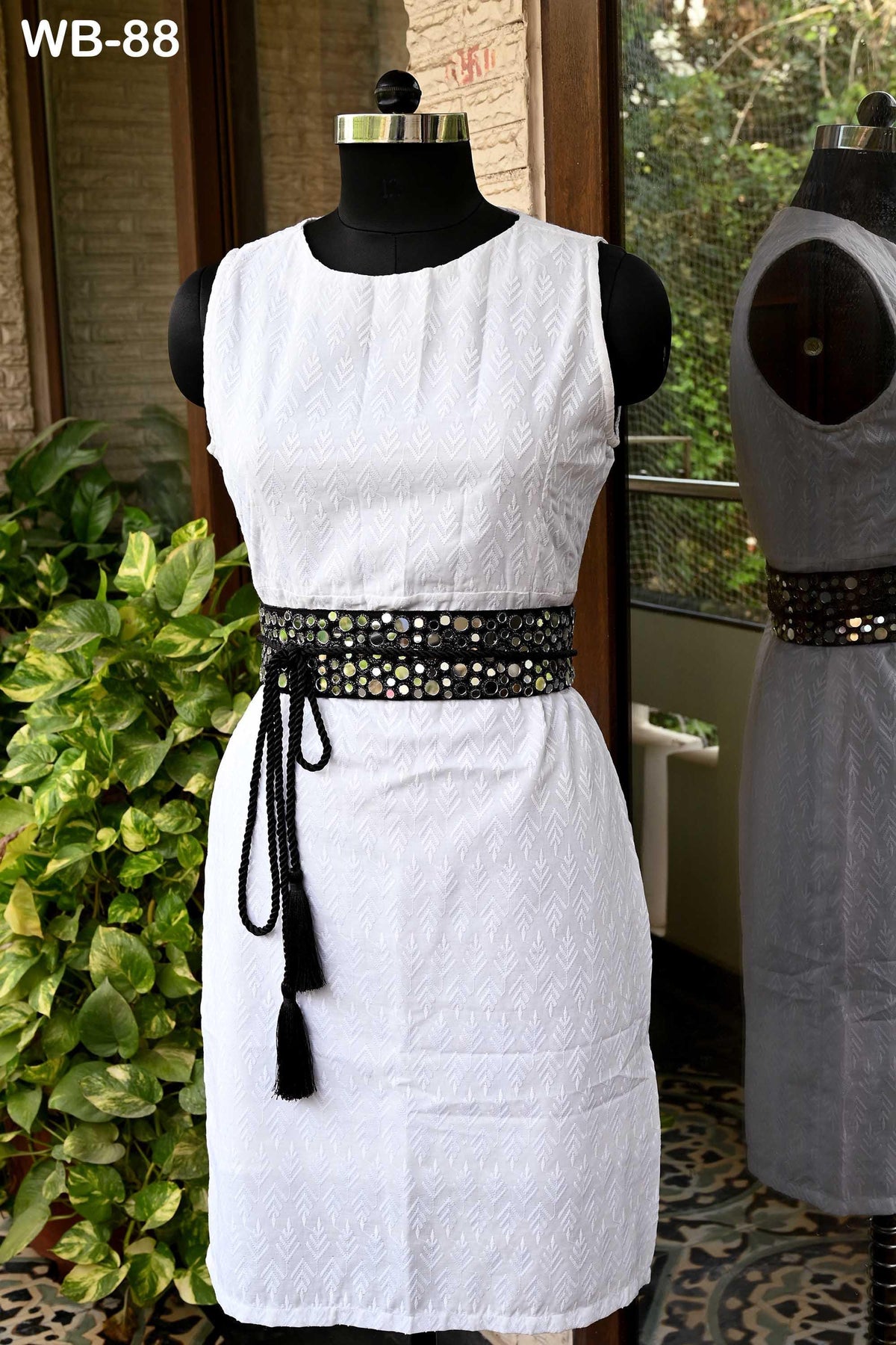 A white sleeveless dress with a black belt featuring a tassel is displayed on a mannequin, with a potted plant in the background.