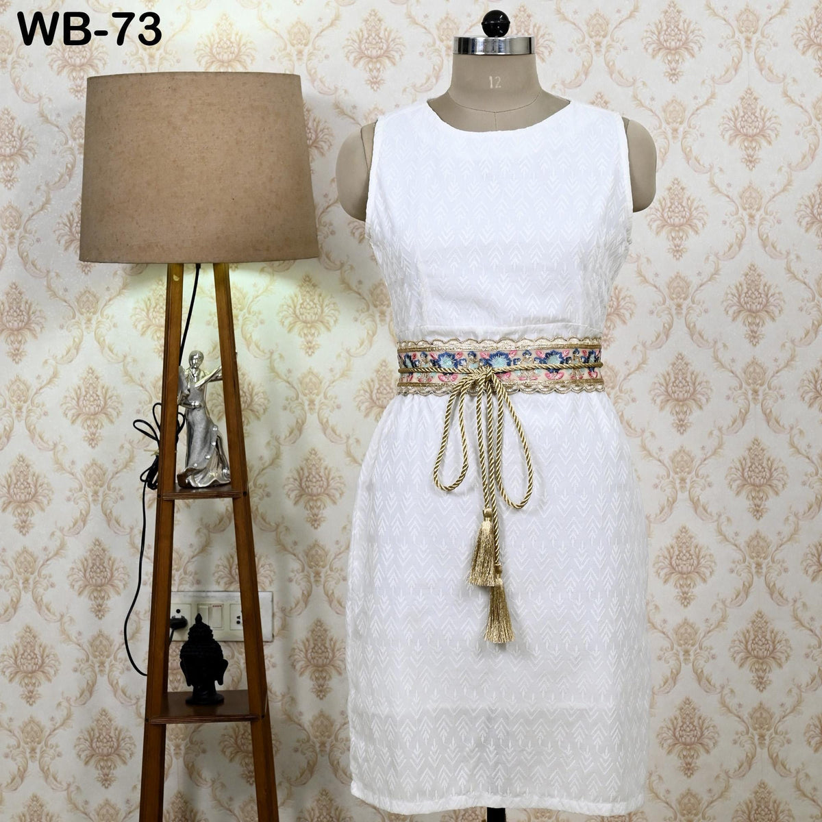 A white dress with a decorative belt is displayed on a mannequin, accompanied by a lamp and a figurine on a wooden stand.