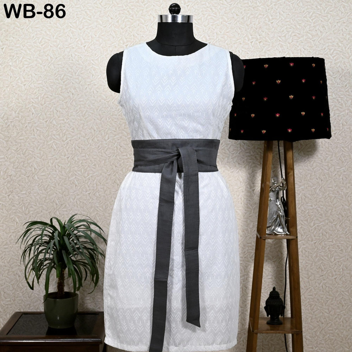 a white sleeveless dress with a gray belt, displayed on a mannequin. The dress is placed next to a black lamp and a potted plant, creating a stylish and elegant setting.