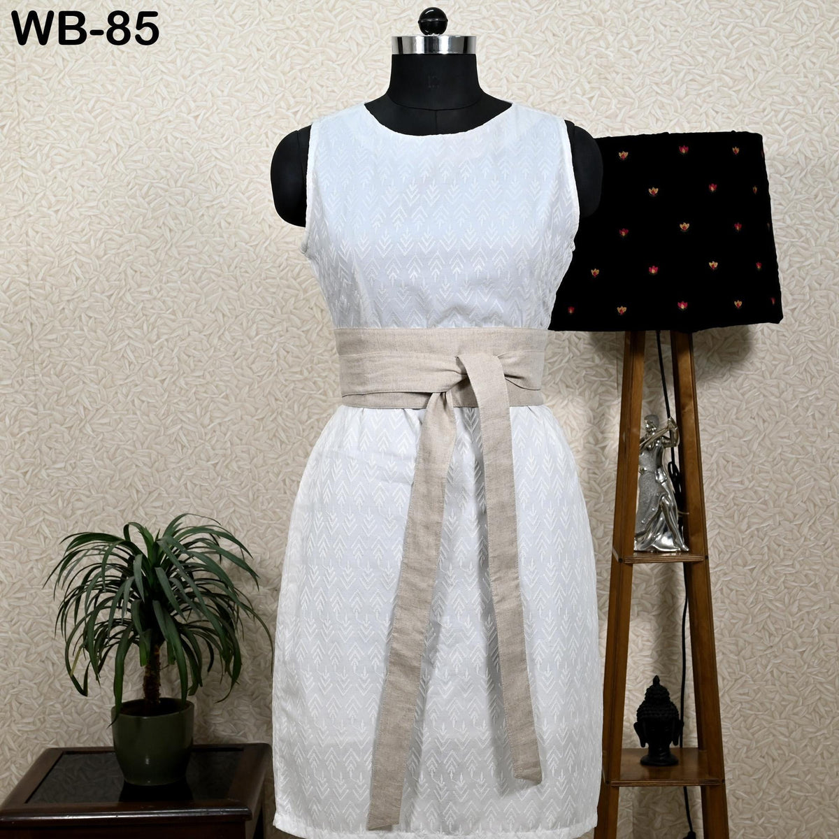 A white sleeveless dress with a beige belt, displayed on a mannequin, with a potted plant and a lamp in the background.