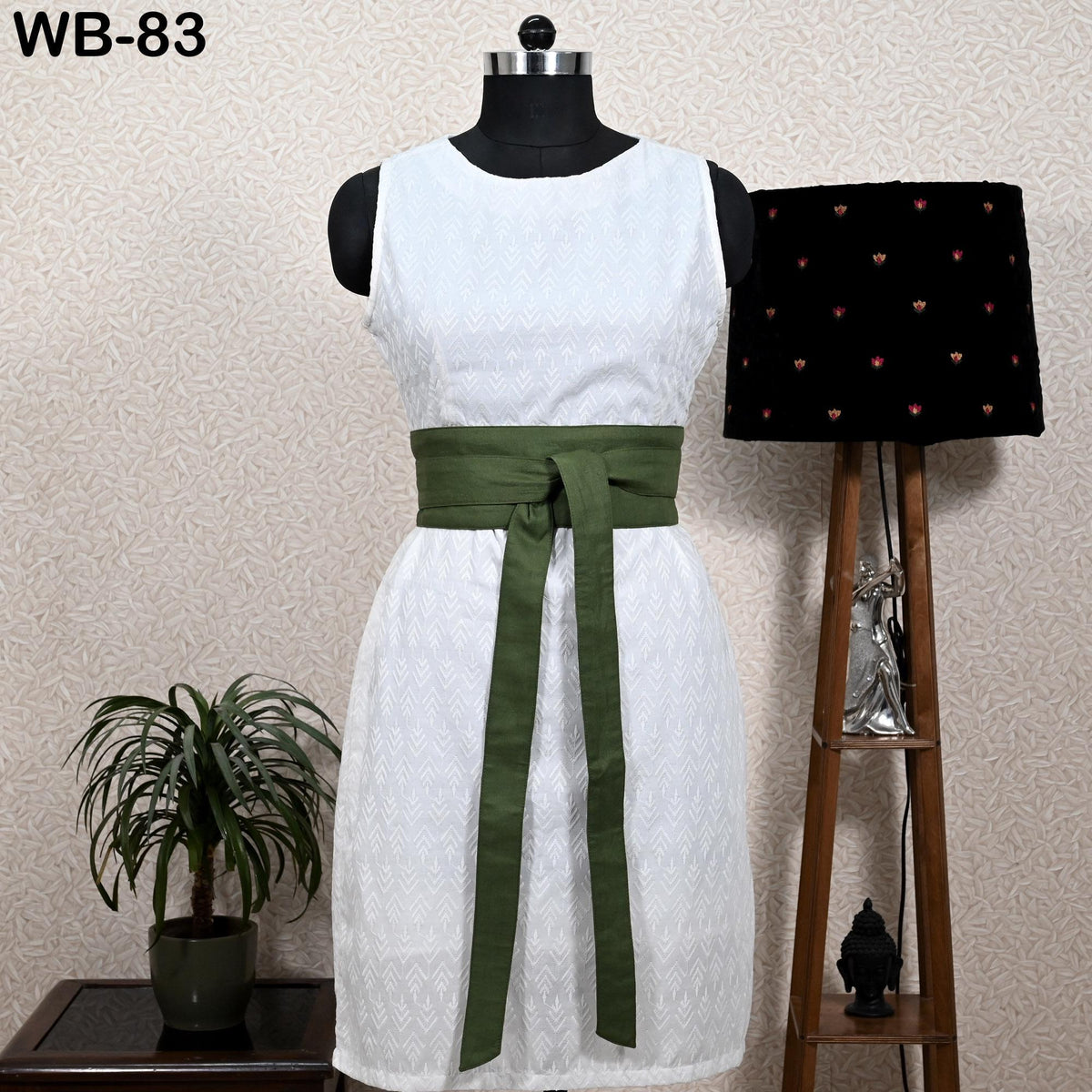 a white dress with a green sash, displayed on a mannequin. The dress is placed on a wooden stand, with a potted plant and a lamp nearby.