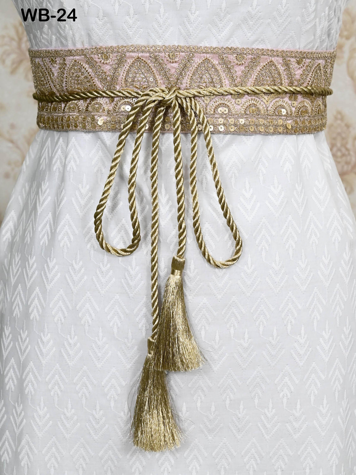 a close-up view of a white dress with a decorative belt featuring a gold and pink pattern. The belt has a tassel at the end and is tied in a bow.