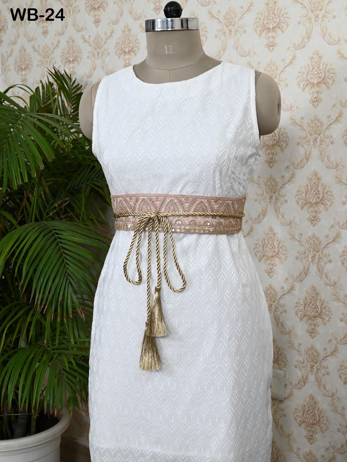 A white sleeveless dress with a decorative belt, displayed on a mannequin against a patterned wallpaper.