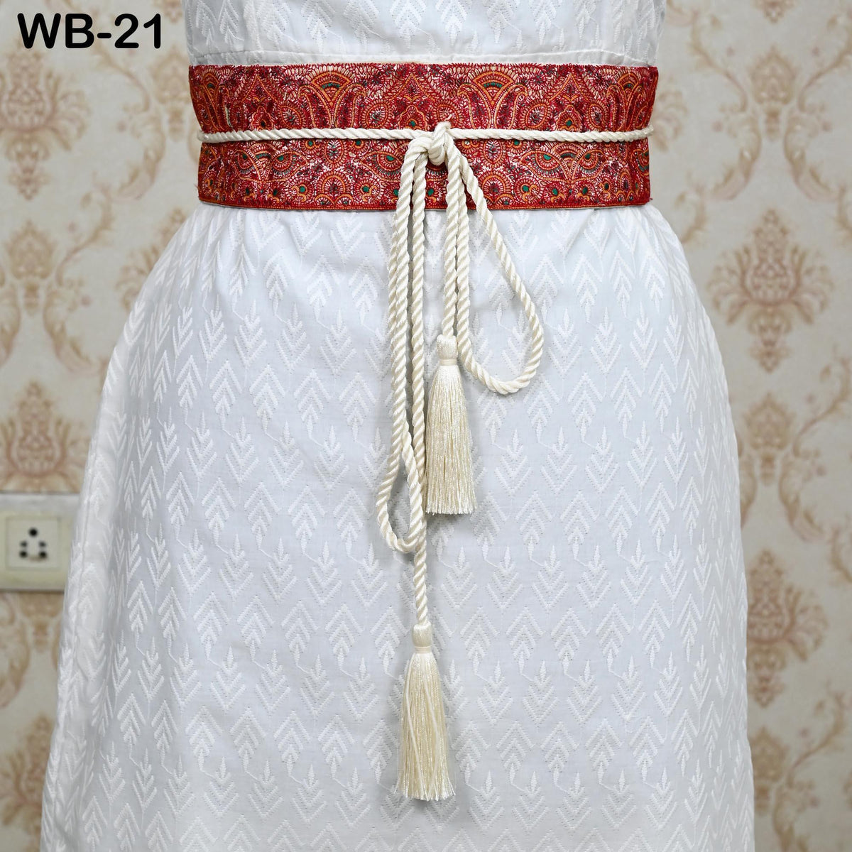 A close-up view of a white dress with a red and white patterned belt, featuring a tassel hanging from the belt.