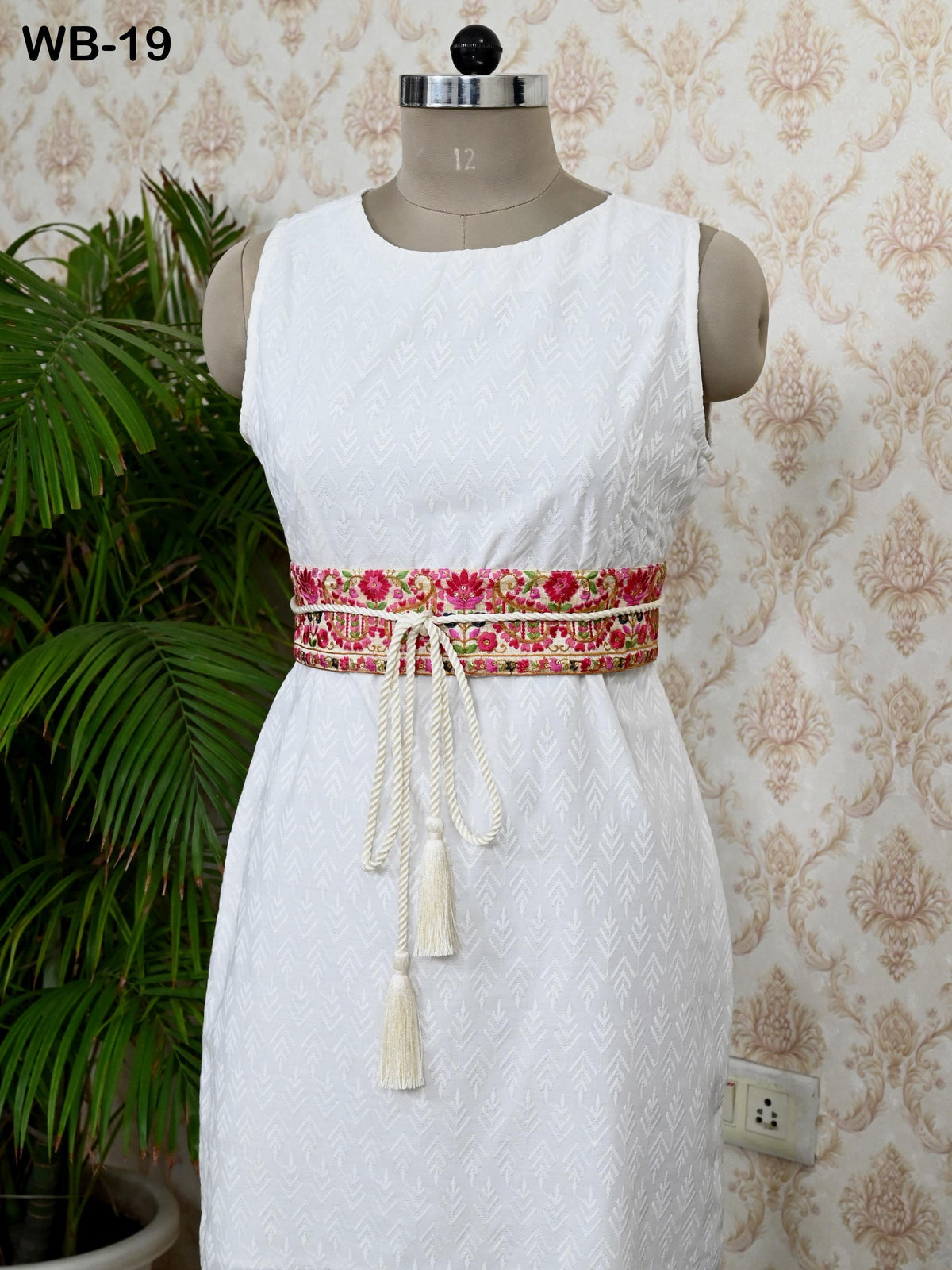 A mannequin wearing a white sleeveless dress with a floral patterned belt, displayed against a patterned wallpaper.