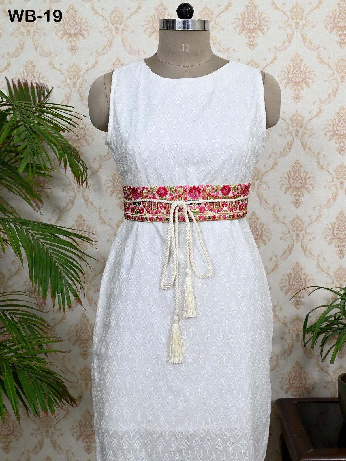 A white dress with a red and pink floral belt displayed on a mannequin against a patterned wallpaper background.