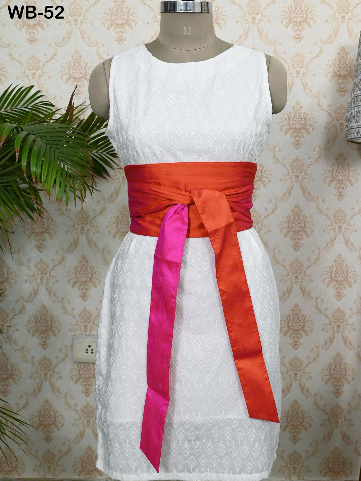 A white dress with a pink and orange sash, displayed on a mannequin against a patterned wallpaper.