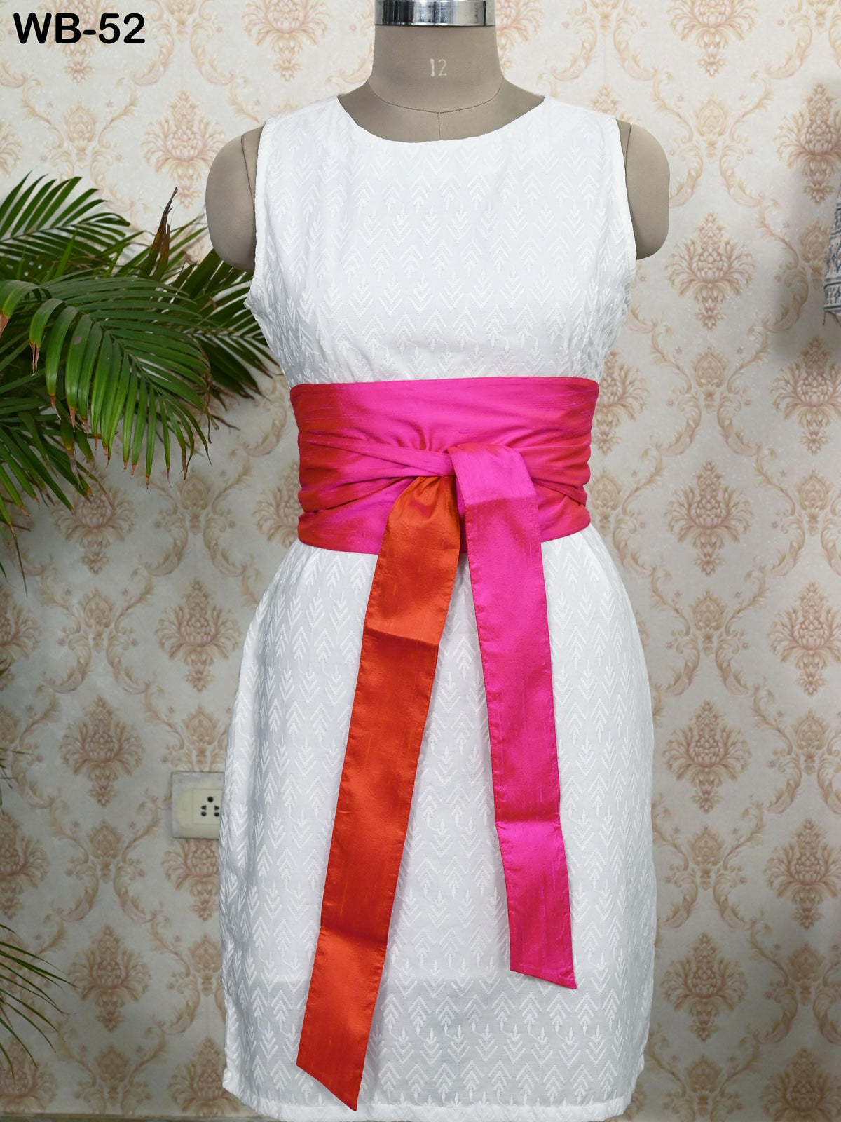 A white dress with a pink sash, displayed on a mannequin against a patterned wallpaper.