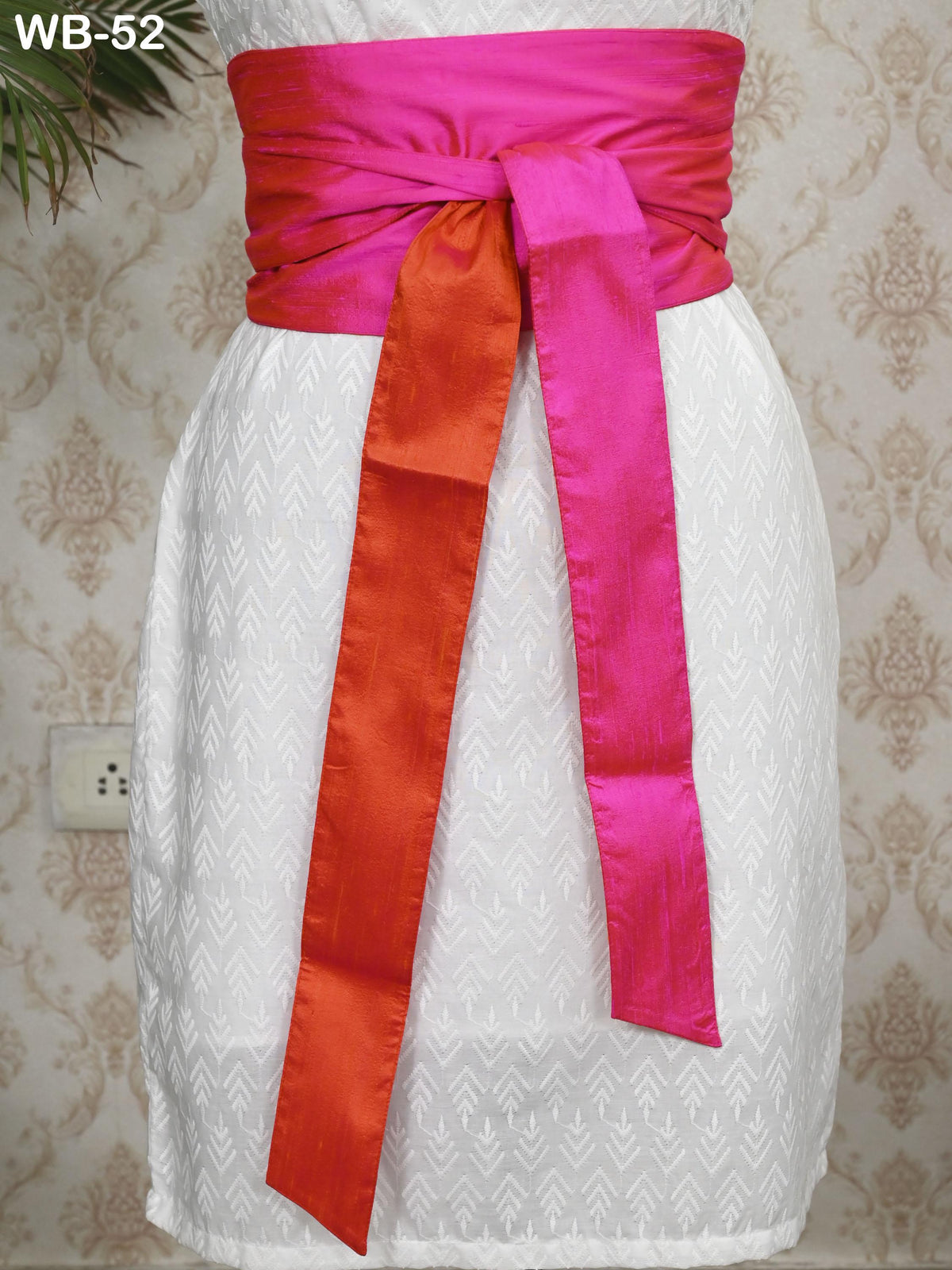 A white dress with a pink sash tied around the waist, displayed against a patterned wall.