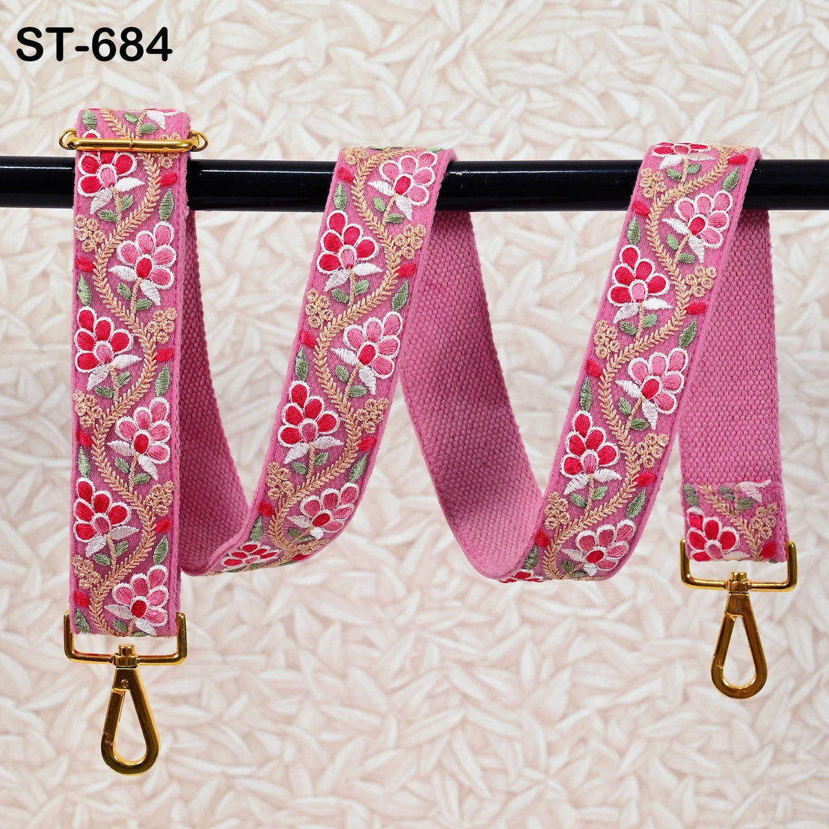 A pink floral embroidered strap with gold hardware, hanging on a black bar.