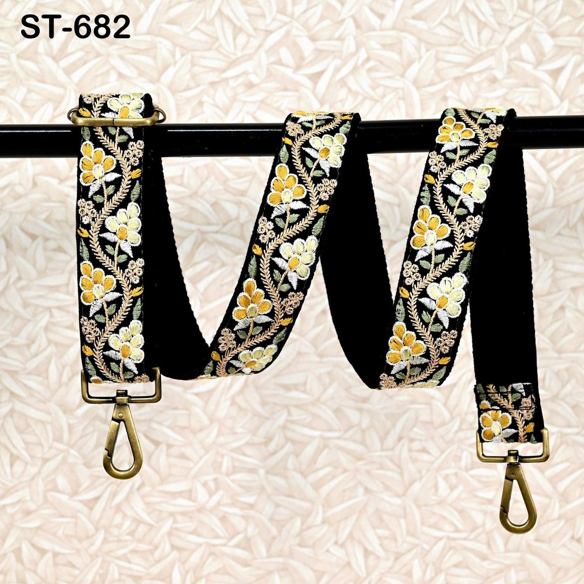 A black and gold floral patterned strap with a gold metal buckle, hanging on a black metal bar.