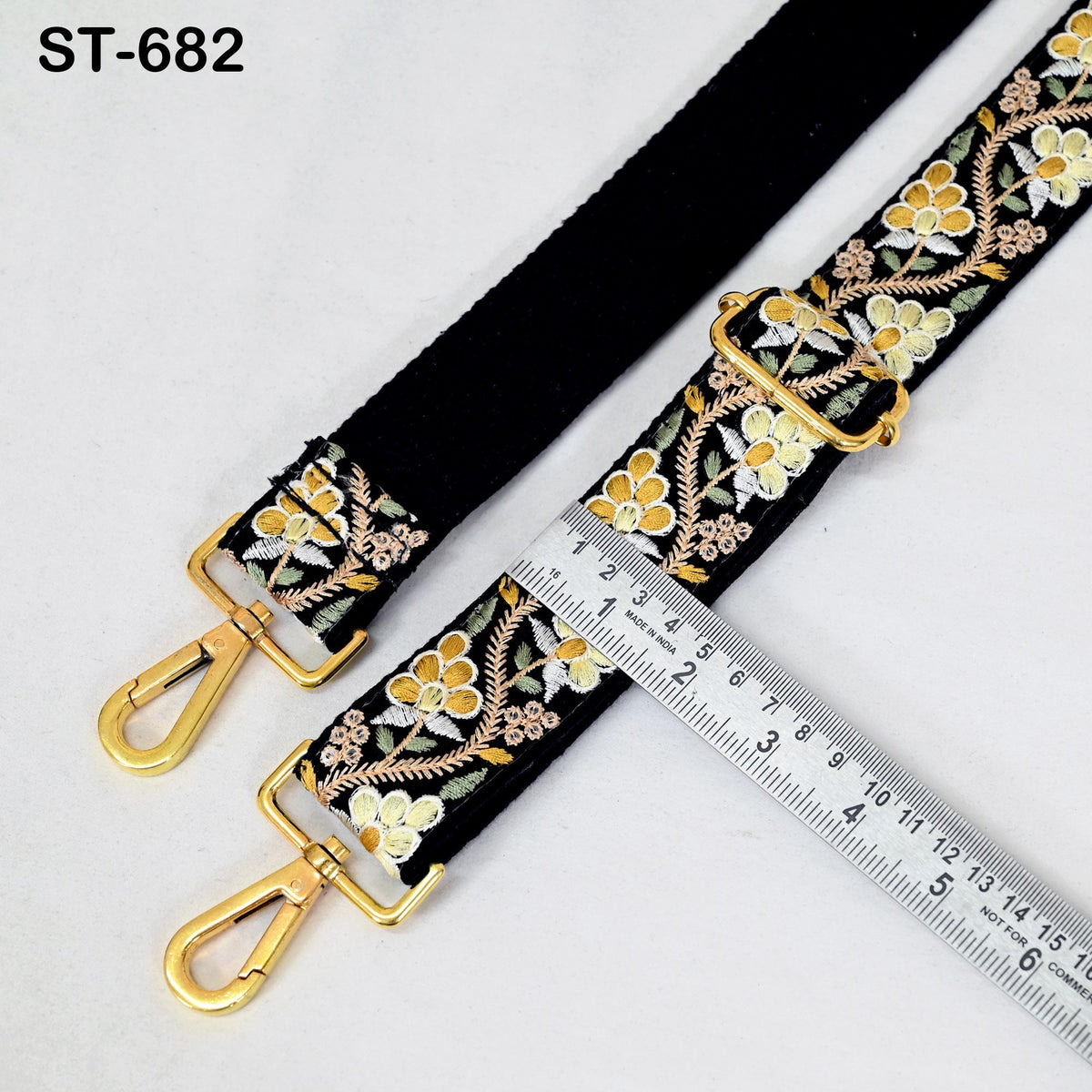 a black strap with a floral pattern and two gold metal hooks, placed next to a ruler for size comparison.