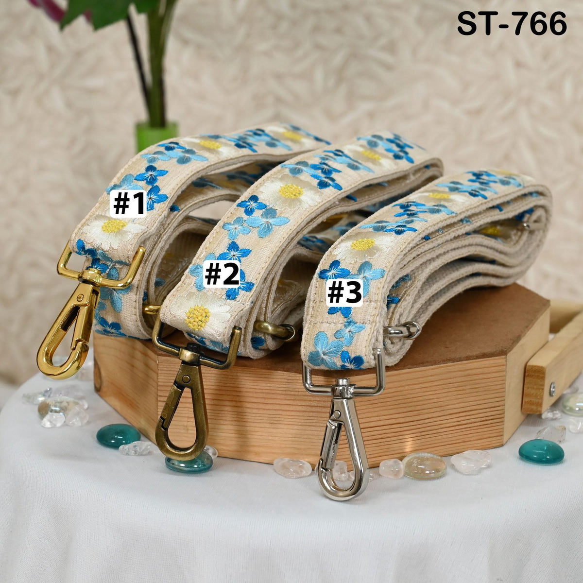 Three floral-patterned fabric straps are displayed on a wooden surface, each with a metal clasp and attached to a gold hook.