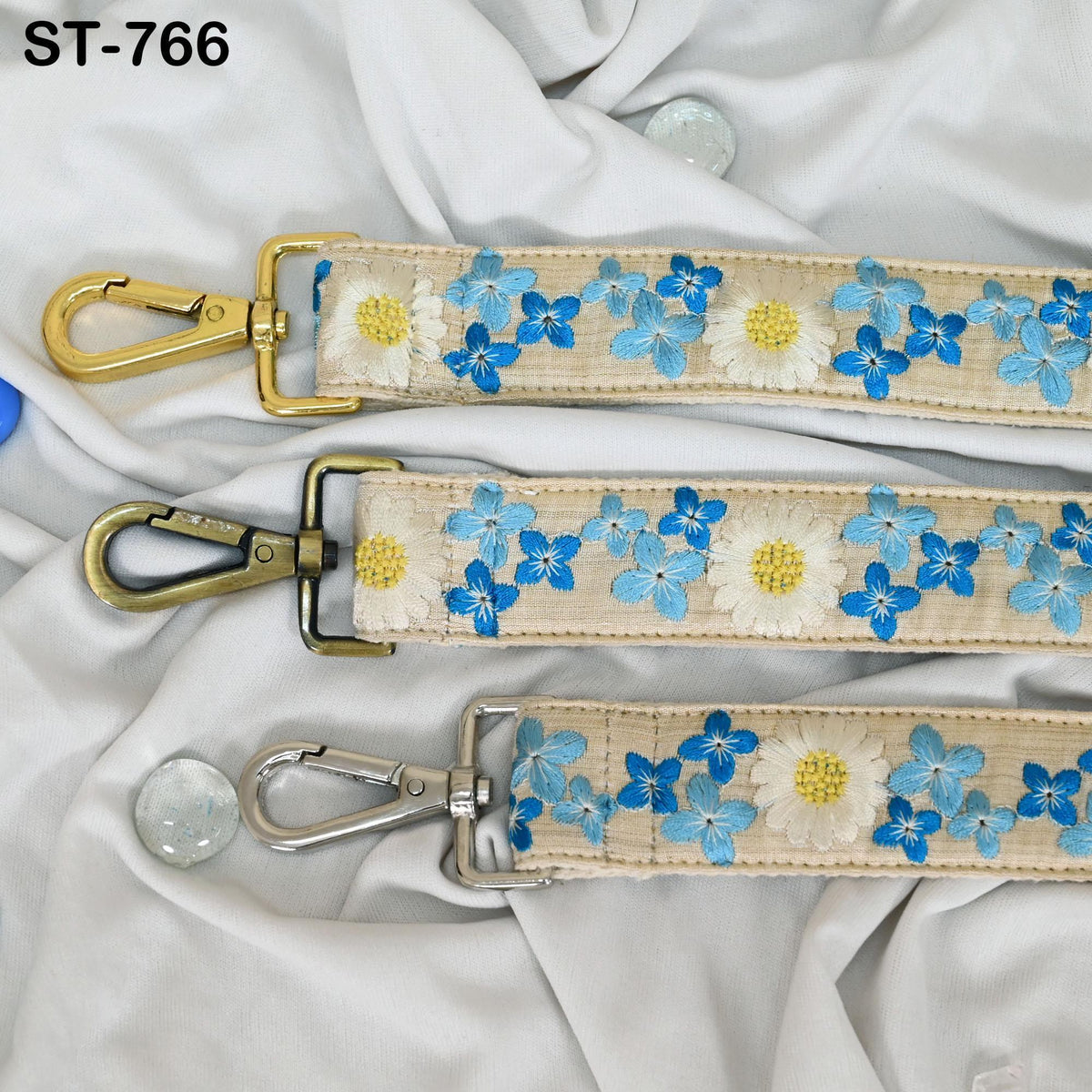 three straps with floral embroidery, each with a metal clasp and hook. The straps are arranged on a white fabric background.