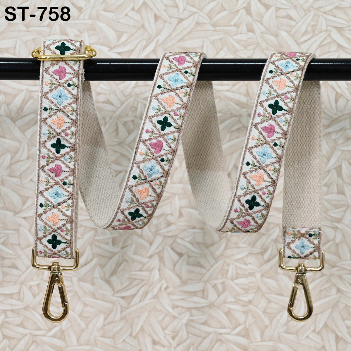 A decorative beige strap with a floral pattern and two metal hooks attached to it, hanging on a black bar.