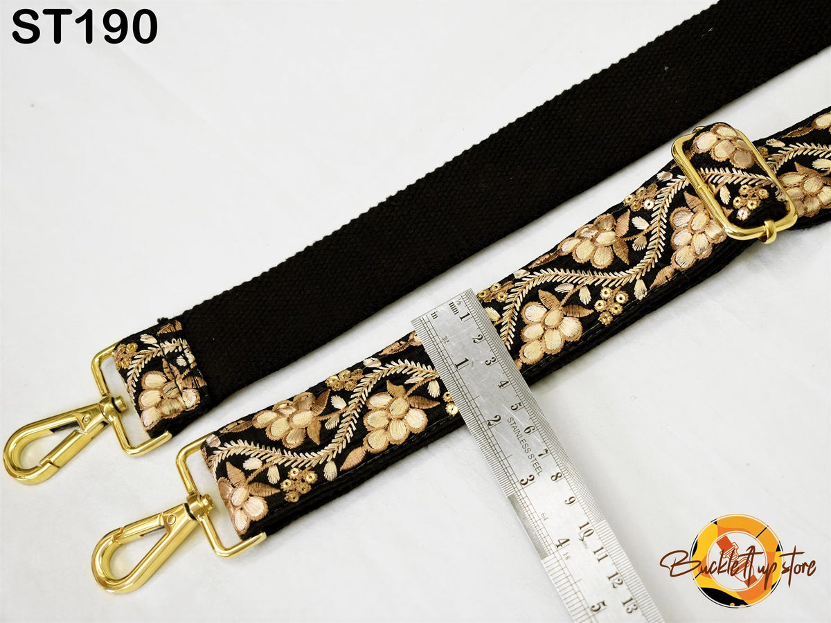 a black strap with a floral pattern and two gold-colored metal hooks, placed next to a ruler for size comparison.