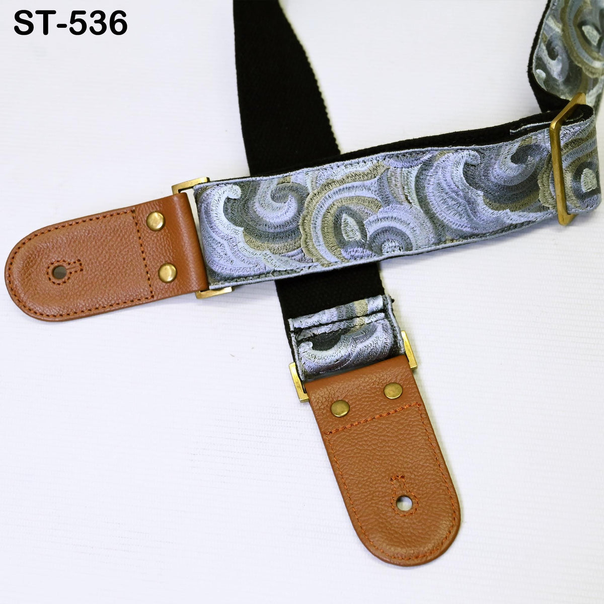 A brown and silver guitar strap with a black and white patterned design, featuring two leather straps with brass buckles.