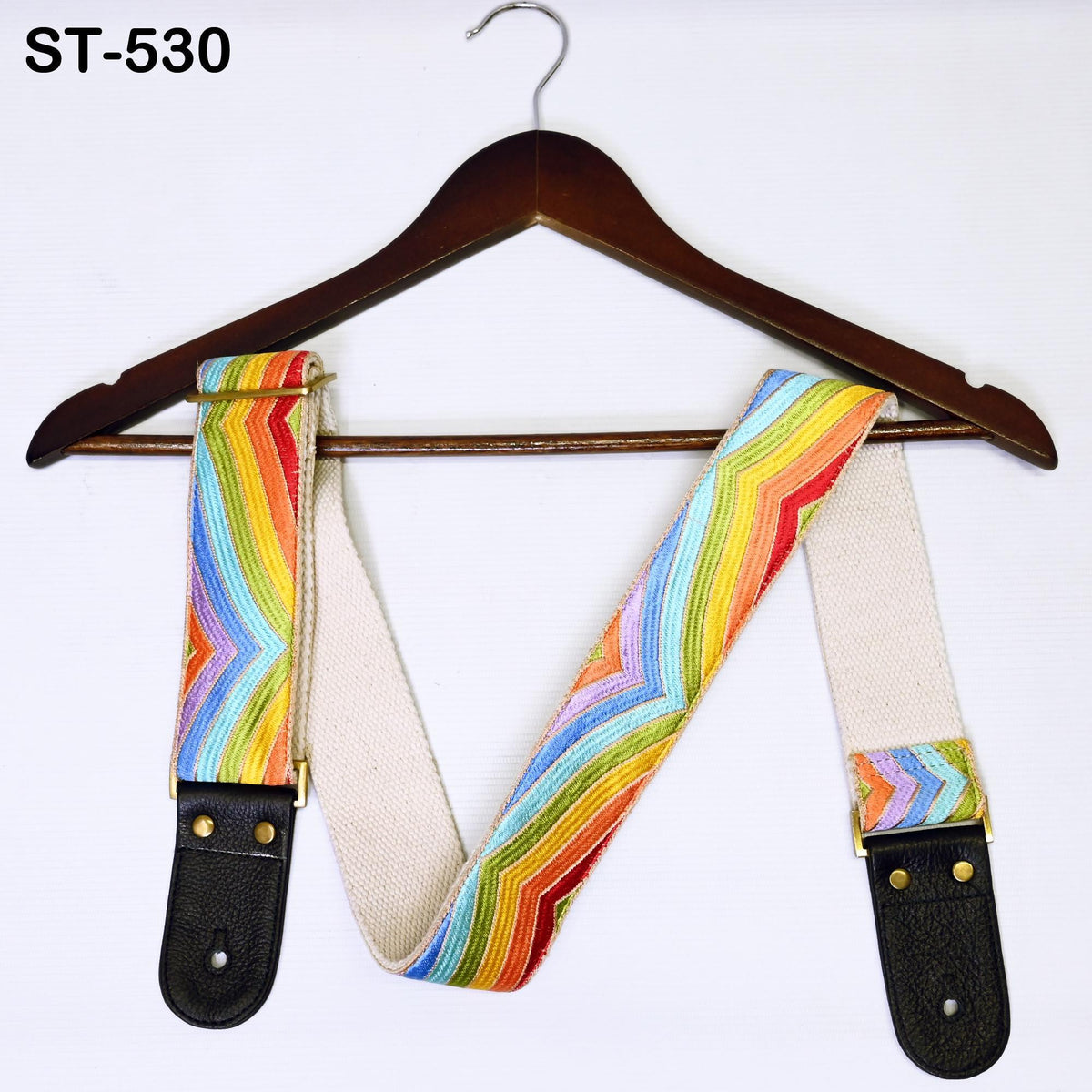 A colorful guitar strap with a rainbow pattern is hanging on a wooden hanger against a white background.