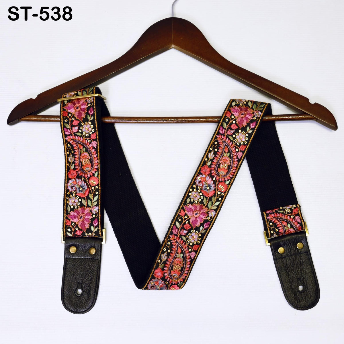 A pair of black guitar straps with a floral pattern and gold accents, hanging on a wooden hanger.