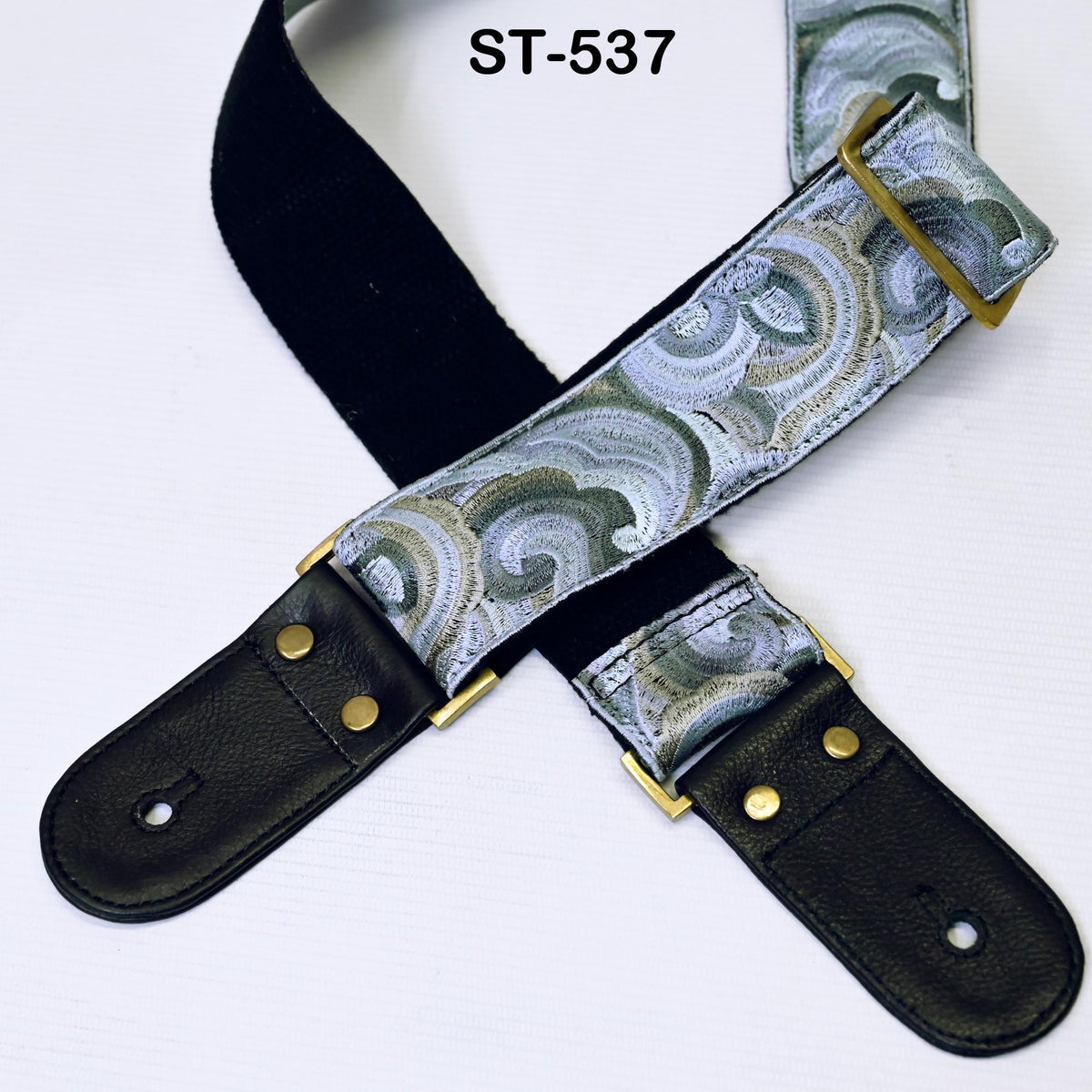 A black and silver guitar strap with a patterned design, featuring gold-colored hardware.