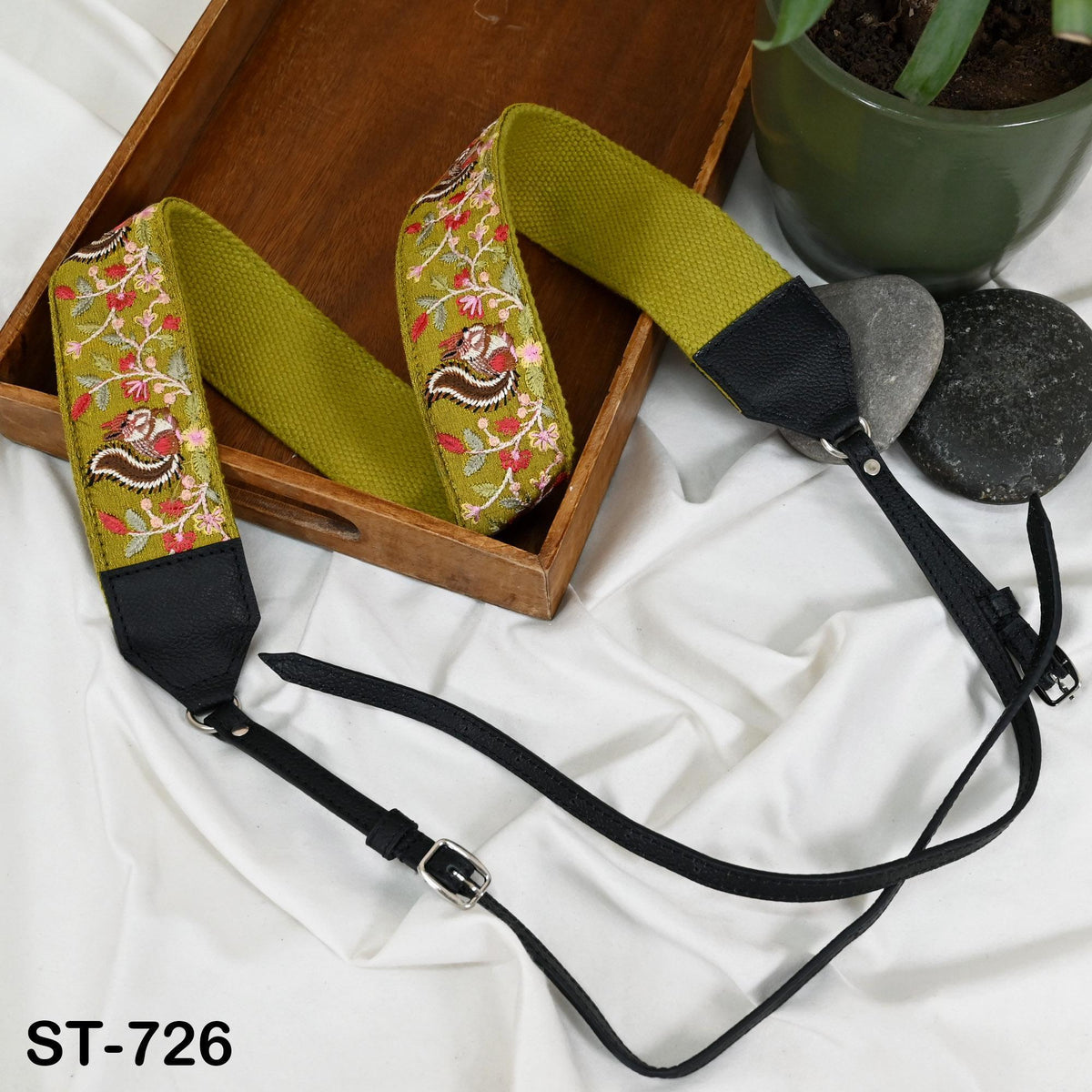 a pair of green and black camera straps with floral embroidery, placed on a wooden surface next to a potted plant and some rocks.