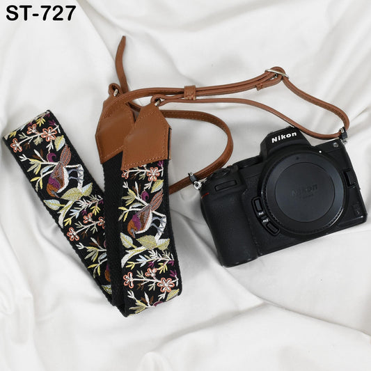 A black and brown camera strap with a floral pattern and a black Nikon camera are placed on a white surface.
