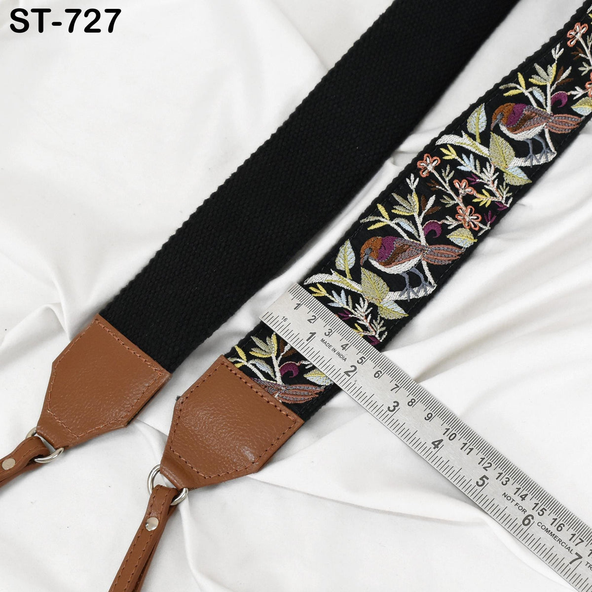 a black strap with a floral embroidered pattern and a brown leather strap, both resting on a white fabric surface. A ruler is placed next to the strap, likely for measuring purposes.