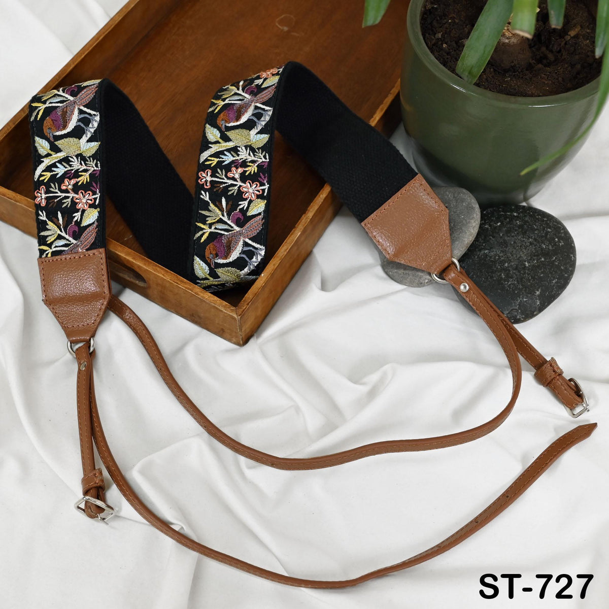 a pair of brown leather camera straps with floral embroidery, placed on a wooden surface next to a potted plant and a rock.