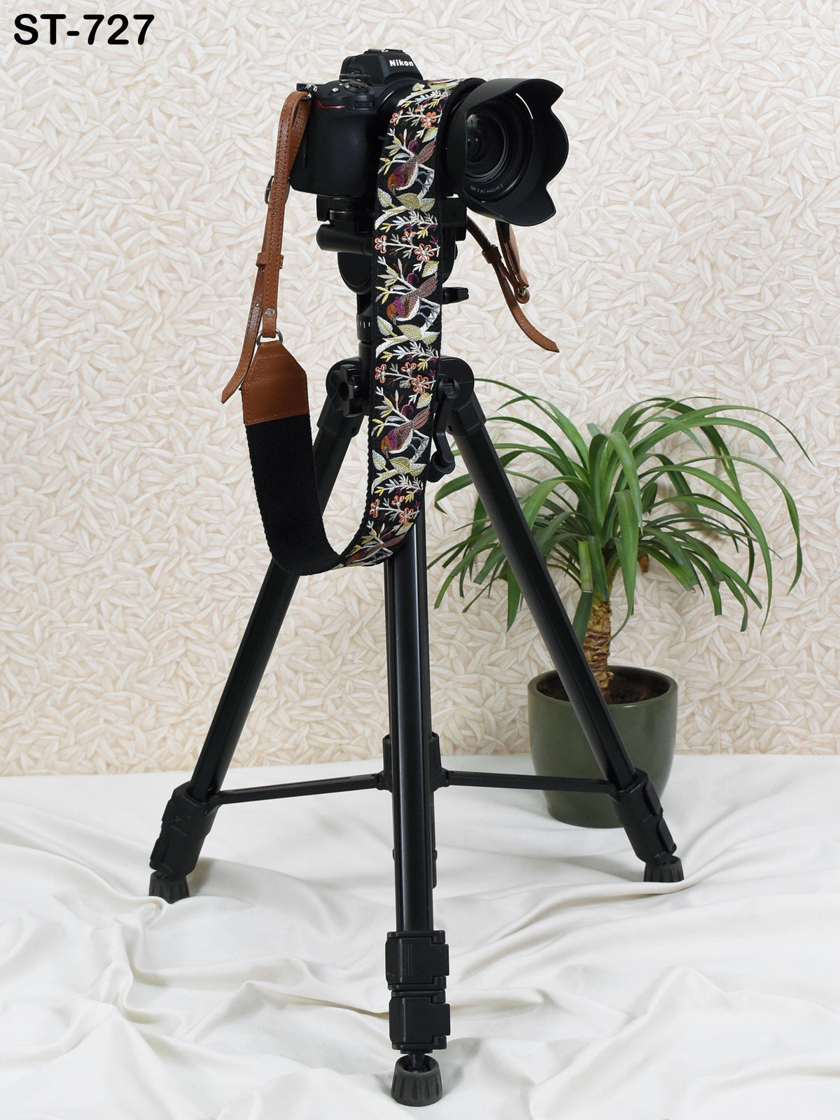 A camera mounted on a tripod, with a patterned strap, is positioned next to a potted plant on a white surface.